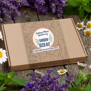 May include: A brown cardboard box with a circular label that reads "Salmon Moon Apothecary GARDEN SEED KIT 10 HERB VARIETIES FOR THE HOME HERBALIST." The box is surrounded by purple lavender, white daisies, and green mint leaves.