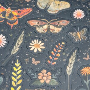 Puede incluir: Pegatinas decorativas con mariposas, flores y follaje en tonos naranja, amarillo y marr&oacute;n sobre un fondo oscuro. Las pegatinas tienen un aspecto texturizado y en relieve, con peque&ntilde;as motas doradas esparcidas.