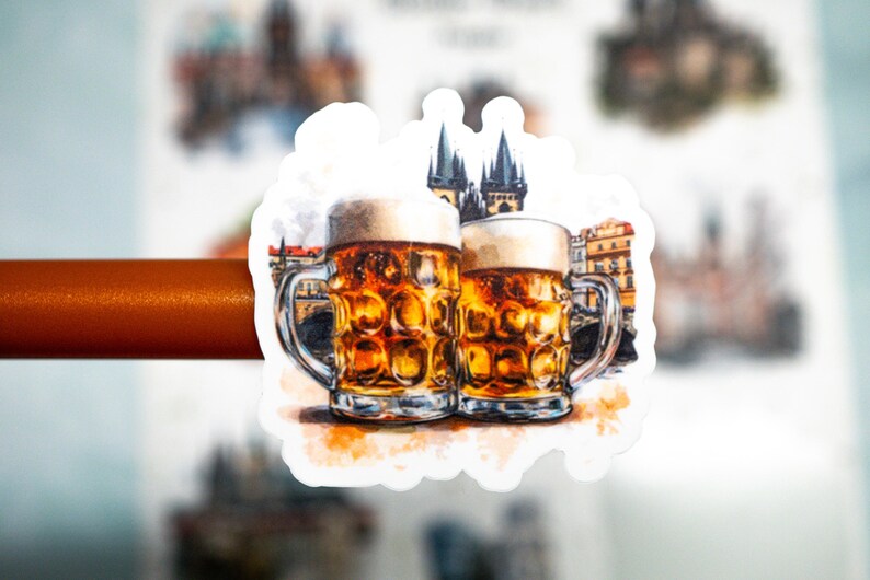 May include: A sticker featuring two beer steins with foamy tops, filled with amber liquid. The background shows a watercolor illustration of a European city with a castle. The steins have silver handles.