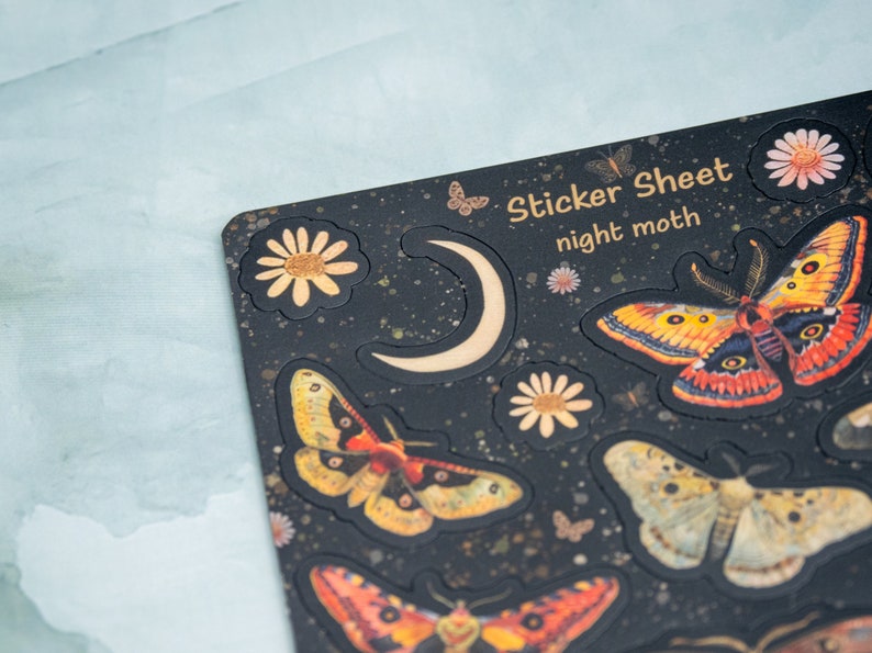 Puede incluir: Una hoja de pegatinas negra con tem&aacute;tica de polilla nocturna. La hoja presenta varias ilustraciones de polillas en tonos naranja, amarillo y marr&oacute;n, junto con una luna creciente y detalles florales. El texto "Sticker Sheet night moth" est&aacute; impreso.