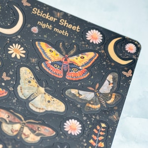 Puede incluir: Una hoja de pegatinas con un fondo oscuro que presenta varios dise&ntilde;os de polillas en naranja, amarillo y marr&oacute;n. La hoja tambi&eacute;n incluye detalles florales y de media luna. El texto en la hoja dice "Sticker Sheet night moth."
