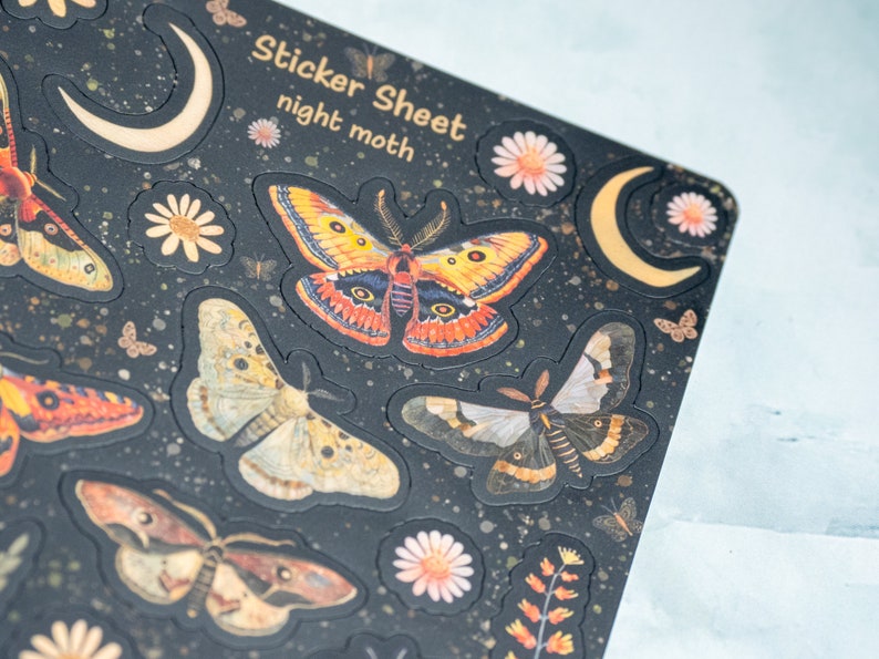 Puede incluir: Una hoja de pegatinas con un fondo oscuro que presenta varios dise&ntilde;os de polillas en naranja, amarillo y marr&oacute;n. La hoja tambi&eacute;n incluye detalles florales y de media luna. El texto en la hoja dice "Sticker Sheet night moth."