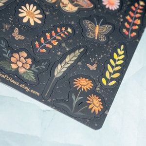 Puede incluir: Una hoja de pegatinas con un fondo oscuro y varios dise&ntilde;os florales e insectos. Las pegatinas presentan mariposas, margaritas y otros elementos bot&aacute;nicos en tonos naranja, amarillo y verde. El texto "HannaCraftHaus.etsy.com" es visible.