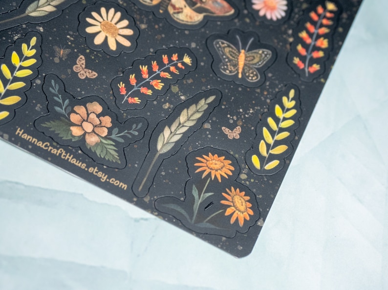 Puede incluir: Una hoja de pegatinas con un fondo oscuro y varios dise&ntilde;os florales e insectos. Las pegatinas presentan mariposas, margaritas y otros elementos bot&aacute;nicos en tonos naranja, amarillo y verde. El texto "HannaCraftHaus.etsy.com" es visible.