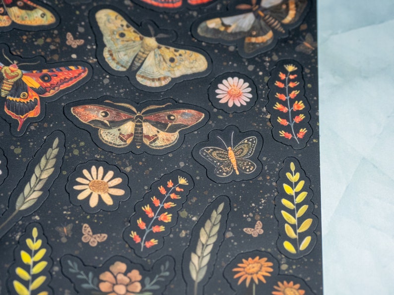 Puede incluir: Hoja de pegatinas con varios dise&ntilde;os de polillas y mariposas, junto con elementos florales y bot&aacute;nicos. Las pegatinas tienen un fondo oscuro con un patr&oacute;n moteado y son en tonos marr&oacute;n, naranja, amarillo y blanco.