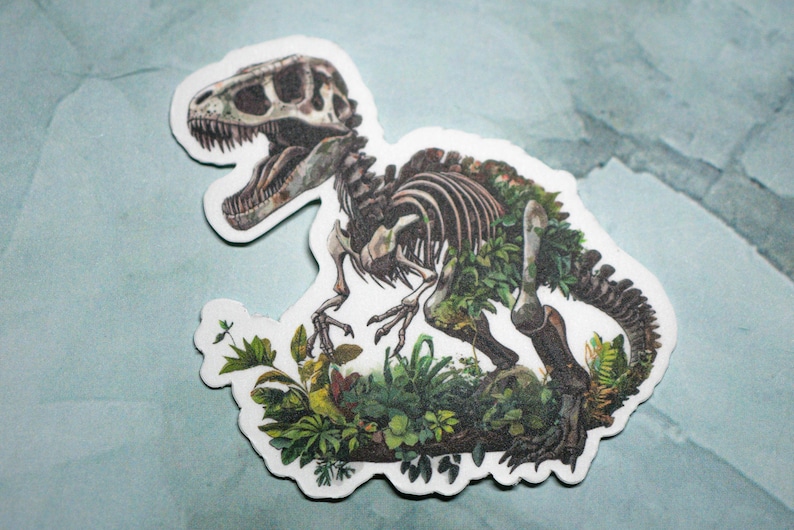 May include: A sticker of a dinosaur skeleton with green foliage growing over the bones. The dinosaur is in a crouching position with its head turned to the side. The sticker is on a light blue background.