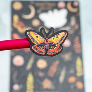 Puede incluir: Una pegatina de mariposa colorida con detalles en naranja, amarillo y negro. La pegatina est&aacute; colocada sobre un bol&iacute;grafo rojo, con un fondo oscuro con dise&ntilde;os florales y celestiales. La mariposa tiene intrincados patrones en las alas.