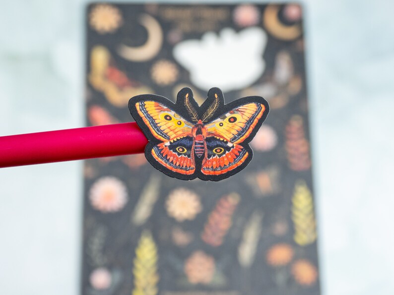 Puede incluir: Una pegatina de mariposa colorida con detalles en naranja, amarillo y negro. La pegatina est&aacute; colocada sobre un bol&iacute;grafo rojo, con un fondo oscuro con dise&ntilde;os florales y celestiales. La mariposa tiene intrincados patrones en las alas.