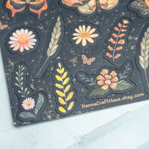 Puede incluir: Hoja de pegatinas decorativas con dise&ntilde;os florales e insectos. Las pegatinas incluyen mariposas, margaritas y espigas de trigo en tonos naranja, amarillo y verde, sobre un fondo oscuro con un patr&oacute;n moteado. El texto "HannaCraftHaus.etsy.com" es visible.