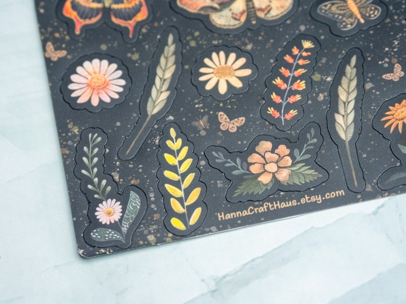 Puede incluir: Hoja de pegatinas decorativas con dise&ntilde;os florales e insectos. Las pegatinas incluyen mariposas, margaritas y espigas de trigo en tonos naranja, amarillo y verde, sobre un fondo oscuro con un patr&oacute;n moteado. El texto "HannaCraftHaus.etsy.com" es visible.