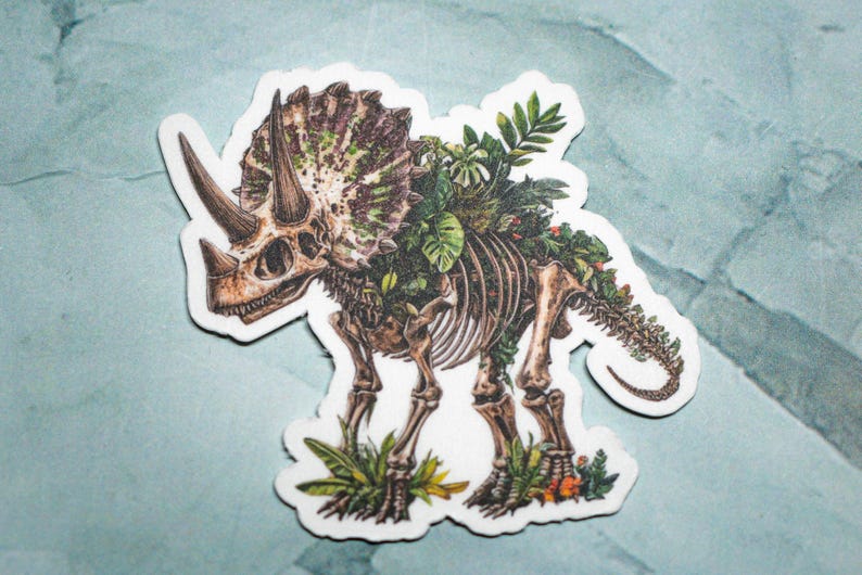 May include: A sticker featuring a detailed illustration of a triceratops skeleton with plants growing over the bones. The sticker is white with a light blue background.