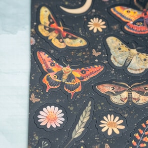 Puede incluir: Hoja de pegatinas con varios dise&ntilde;os de polillas y mariposas en tonos naranja, amarillo y marr&oacute;n, con detalles florales y celestiales. Las pegatinas est&aacute;n sobre un fondo negro con textura.