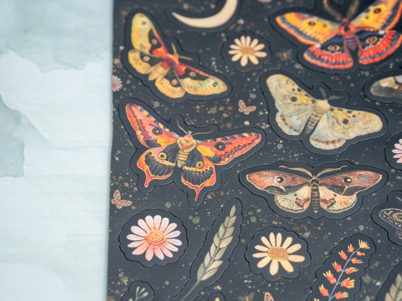 Puede incluir: Hoja de pegatinas con varios dise&ntilde;os de polillas y mariposas en tonos naranja, amarillo y marr&oacute;n, con detalles florales y celestiales. Las pegatinas est&aacute;n sobre un fondo negro con textura.