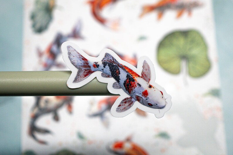 May include: A white, red, and black koi fish sticker with a white background. The sticker is on a gray surface.