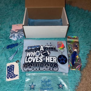 May include: An open cardboard box with a light blue t-shirt that reads "Just a girl who loves her Cowboys." The box contains a scrunchie, a keychain, a pen, and other accessories. The items are on a blue shag rug.