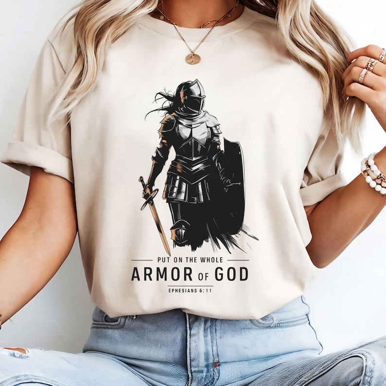 Op de afbeelding: Een beige T-shirt met een zwart-witte illustratie van een vrouw in harnas die een zwaard en schild vasthoudt. De tekst "Trek de volledige wapenrusting van God aan Efezi&euml;rs 6:11" staat op het T-shirt gedrukt.