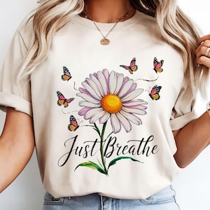 May include: Beige t-shirt with a daisy design. The daisy has white petals, a yellow centre, and a green stem. Butterflies in shades of orange, pink, and black surround the flower. The words "Just Breathe" are written below.