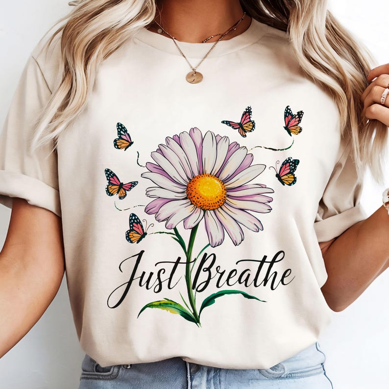 May include: Beige t-shirt with a daisy design. The daisy has white petals, a yellow centre, and a green stem. Butterflies in shades of orange, pink, and black surround the flower. The words "Just Breathe" are written below.