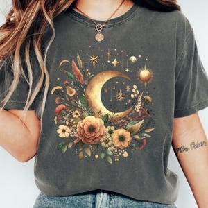 May include: A gray t-shirt with a gold crescent moon and floral design. The moon is surrounded by stars and a sun. The flowers are in shades of orange, yellow, and white.