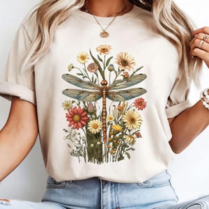 K&ouml;nnte beinhalten: Ein beiges T-Shirt mit einem grafischen Druck einer Libelle, umgeben von Blumen in Gelb-, Orange-, Rot- und Blaut&ouml;nen.