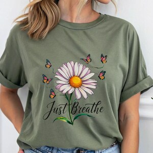 May include: An olive green t-shirt with a daisy design. The daisy has a yellow centre and white petals, with butterflies around it. The words "Just Breathe" are written below the flower. A relaxed, nature-inspired design.