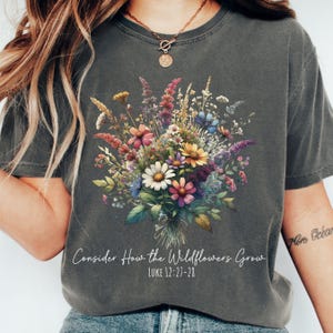 Puede incluir: Una camiseta gris con un diseño floral y el texto "Consider How the Wildflowers Grow" y "LUKE 12:27-28".