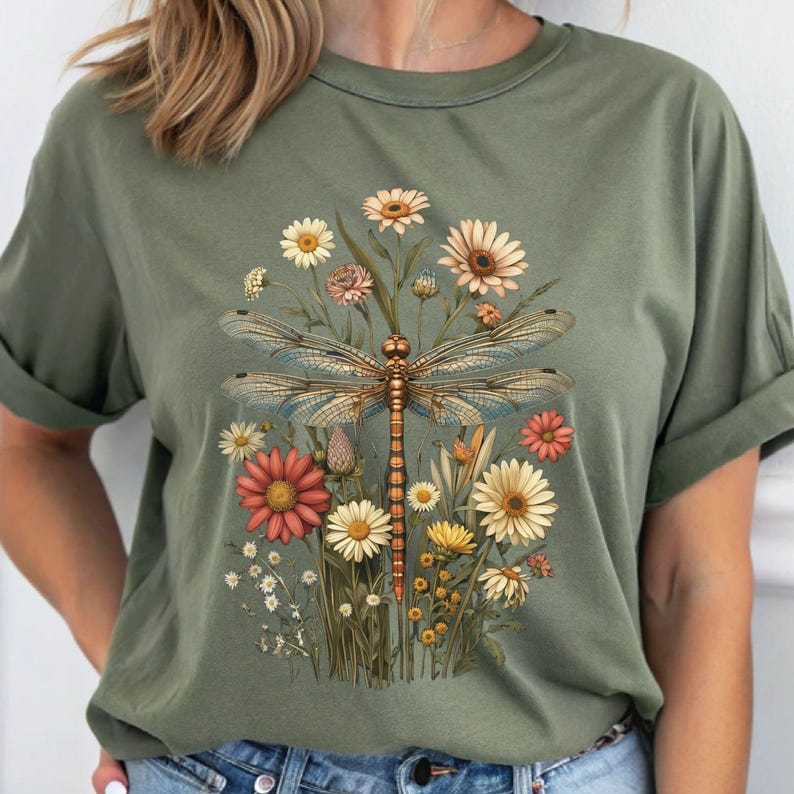 K&ouml;nnte beinhalten: Ein gr&uuml;nes T-Shirt mit einem floralen Druck, der eine Libelle und verschiedene Blumen in Rosa-, Gelb- und Wei&szlig;t&ouml;nen zeigt.