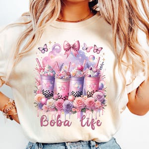 Puede incluir: Una camiseta blanca con un gráfico de cuatro vasos de té de boba con acentos rosas, morados y azules. Los vasos están rodeados de flores y mariposas. El texto "Boba Life" está impreso debajo de los vasos.