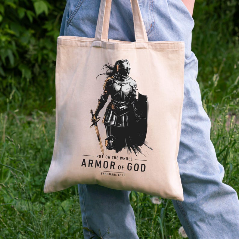 Op de afbeelding: Een beige tote bag met een zwart-witte illustratie van een vrouw in harnas met een zwaard en schild. De tekst "Trek de volledige wapenrusting van God aan Efezi&euml;rs 6:11" staat op de tas gedrukt.