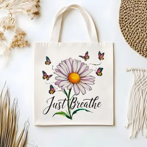 May include: Cream tote bag with a large daisy design, featuring a yellow centre and purple petals, and butterflies. The words "Just Breathe" are written below the flower. The bag has cream handles. The bag is made of a cream-colored material.