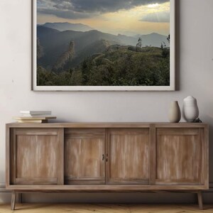 May include: A framed photograph of a mountain range with a cloudy sky and a sunbeam shining through the clouds. The mountains are covered in green vegetation.