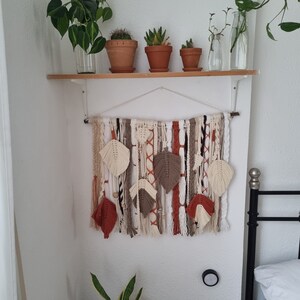 Peut inclure: Une suspension murale en macramé avec du fil brun, blanc et orange. La suspension présente une série de plumes tissées et est exposée sur un mur blanc avec une étagère au-dessus. Il y a plusieurs plantes en pot sur l'étagère.