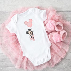 May include: White baby bodysuit with a Mickey Mouse cartoon holding a pink balloon. The outfit includes pink baby shoes with flower details, arranged on a pink tulle skirt. A delightful baby outfit.