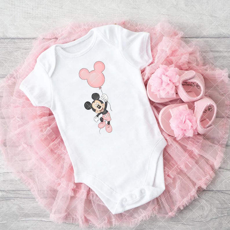 May include: White baby bodysuit with a Mickey Mouse cartoon holding a pink balloon. The outfit includes pink baby shoes with flower details, arranged on a pink tulle skirt. A delightful baby outfit.