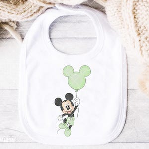 May include: A white baby bib with a cartoon illustration of Mickey Mouse holding a green balloon. Mickey is in black, white, and green, with a happy expression. The bib has a rounded shape and a neck opening for easy use.