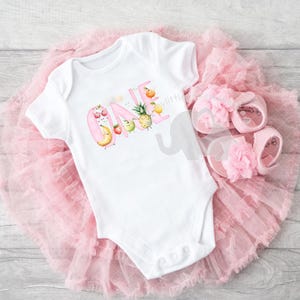 Peut inclure: Un body blanc avec un 'UN' rose orthographi&eacute; avec des illustrations de fruits. Le body est associ&eacute; &agrave; un tutu en tulle rose et des sandales roses.