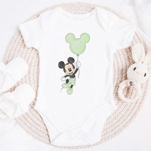 May include: White baby bodysuit with a Mickey Mouse cartoon holding a green balloon. The bodysuit is next to white baby booties and a knitted bunny rattle. The design features green, black, and white colours.