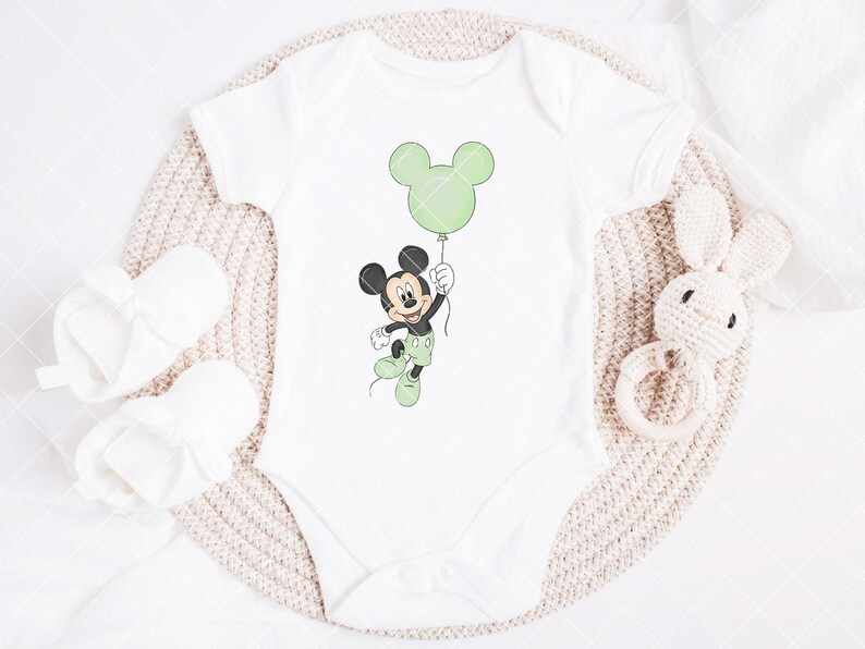 May include: White baby bodysuit with a Mickey Mouse cartoon holding a green balloon. The bodysuit is next to white baby booties and a knitted bunny rattle. The design features green, black, and white colours.