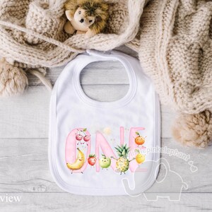Peut inclure: Bavoir rose avec le mot "ONE" en lettres roses entour&eacute; de fruits et de fleurs. Les fruits comprennent une banane, un ananas, des fraises et des cerises.