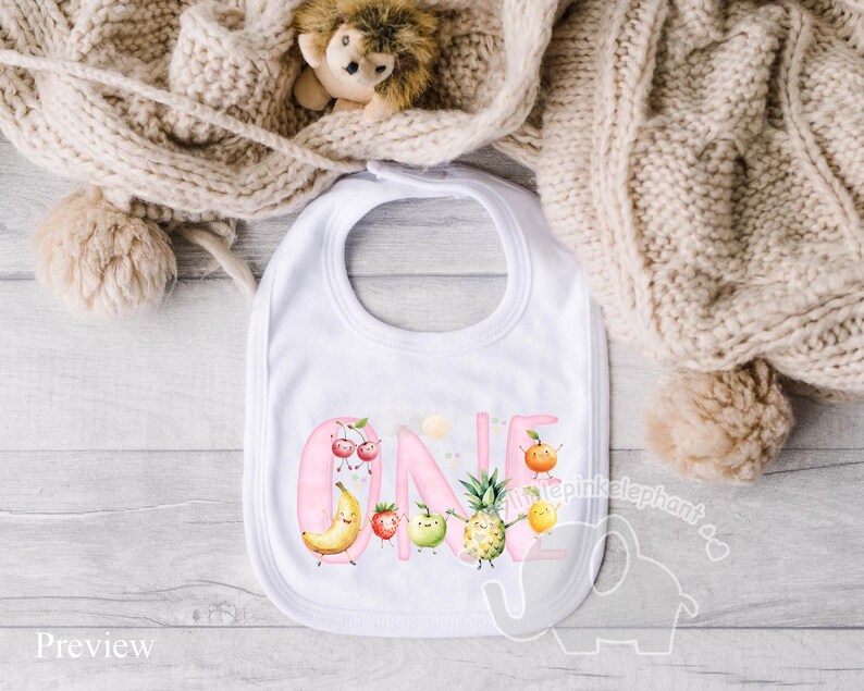 Peut inclure: Bavoir rose avec le mot "ONE" en lettres roses entour&eacute; de fruits et de fleurs. Les fruits comprennent une banane, un ananas, des fraises et des cerises.