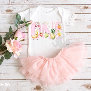 Peut inclure: Un body b&eacute;b&eacute; rose et blanc avec un motif de fruits et le mot "ONE" en lettres roses. Le body est associ&eacute; &agrave; un tutu en tulle rose.