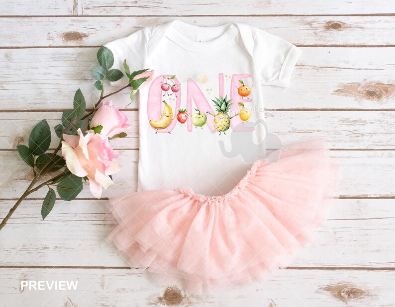 Peut inclure: Un body b&eacute;b&eacute; rose et blanc avec un motif de fruits et le mot "ONE" en lettres roses. Le body est associ&eacute; &agrave; un tutu en tulle rose.