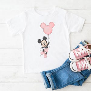 May include: White t-shirt with a Mickey Mouse cartoon holding a pink balloon. The image includes blue denim jeans and pink canvas trainers. A cheerful design for children.