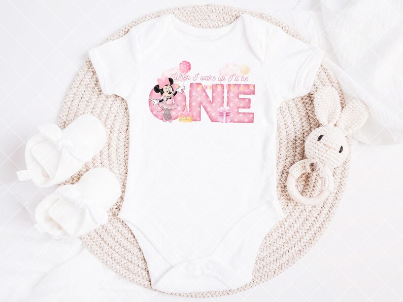 Puede incluir: Body de beb&eacute; blanco con el texto rosa "When I wake up I'll be ONE". El dise&ntilde;o incluye un gr&aacute;fico de Minnie Mouse. Tambi&eacute;n hay un par de patucos blancos y un juguete de conejo de ganchillo.