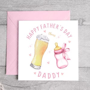 May include: A white card with pink text that reads "Happy Father's Day Daddy" with a glass of beer and a baby bottle with a pink bow. The text "Cheers" is written in pink.