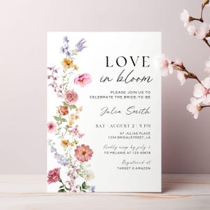 May include: A white invitation card with the text "LOVE in bloom" and floral accents. The card includes details for a bridal celebration, with the bride's name, date, time, and location. RSVP information is also provided.
