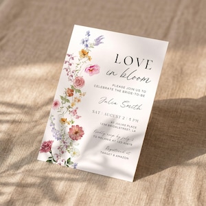 May include: A white invitation card with the text "LOVE in bloom" and floral accents. The card includes details for a bridal celebration, with the bride's name, date, time, and location. The card is decorated with a colorful floral border.