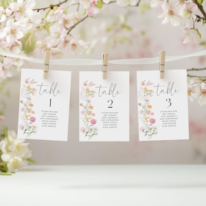 May include: Three white table cards, each with the word "table" in script and a floral border. The cards are numbered 1, 2, and 3, and are held by wooden pegs. The cards are decorated with pastel flowers.