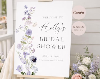 May include: A white rectangular bridal shower welcome sign on a wooden easel featuring a vertical floral illustration of purple and blue flowers on the left side. The text reads: Welcome to Holly's Bridal Shower, April 19, 2025, 15 days until she says I do.