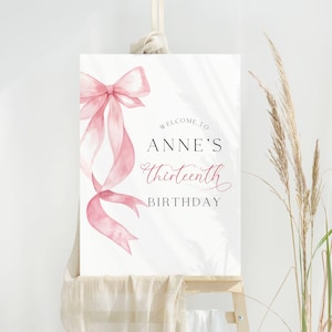 May include: A white sign featuring a pink watercolour bow and ribbons. The text reads "Welcome to Anne's Thirteenth Birthday" in a stylish script and serif font. The sign is displayed on a wooden easel.